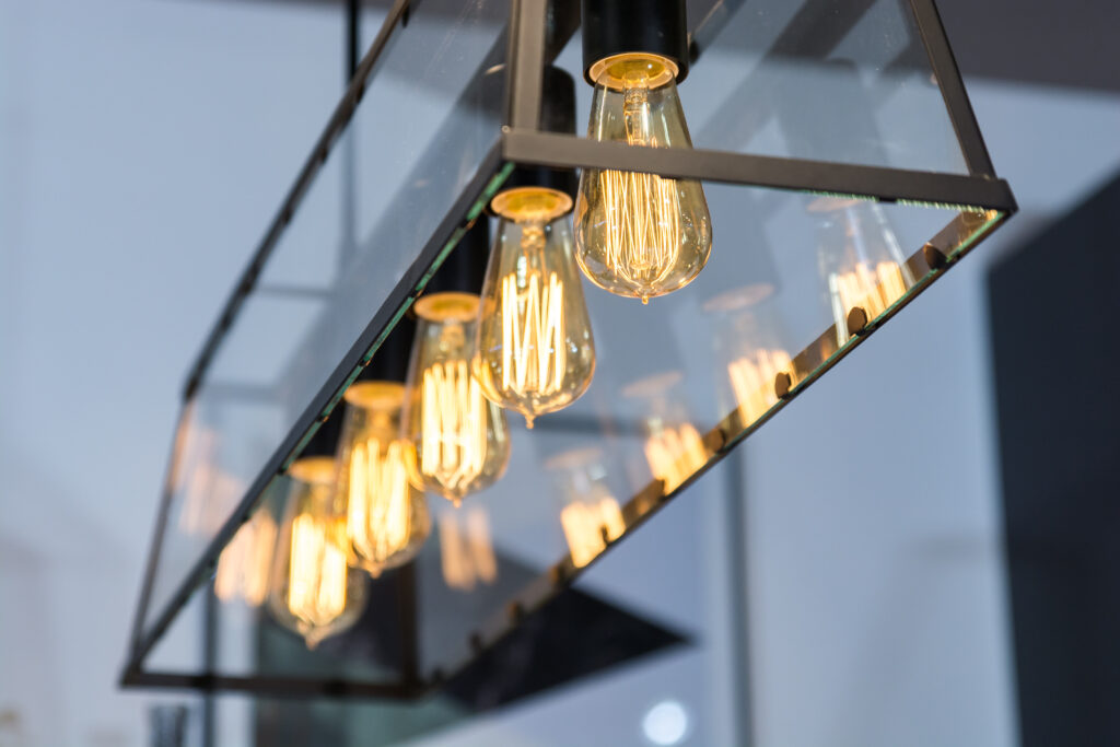 Close-up of a modern rectangular glass and metal light fixture with exposed vintage-style Edison bulbs glowing warmly, reflecting off the glass panels.