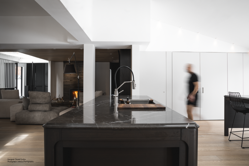 A modern kitchen with a dark marble island and sleek fixtures, open to a living area with neutral sofas and a fireplace. A blurred person walks through the bright, minimalist kitchen space with white walls and wooden floors.