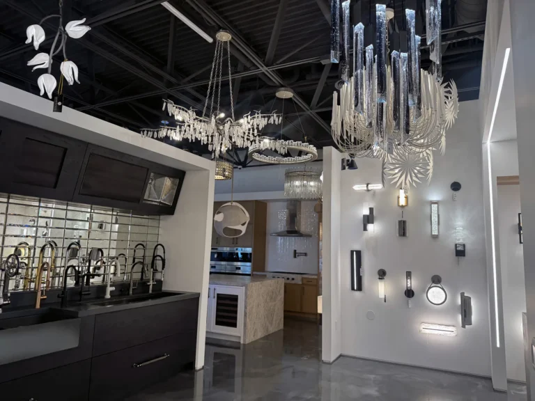 A modern showroom with various contemporary chandeliers and wall lights on display, along with kitchen faucets mounted on a counter and sleek cabinetry under bright, reflective lighting.