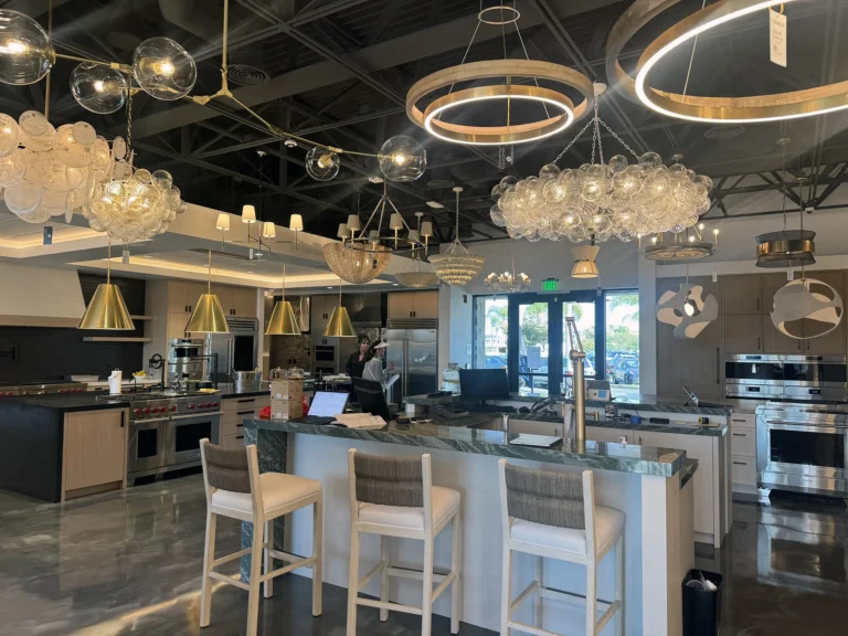 A modern showroom displays various kitchen appliances and lighting fixtures. There is an island with barstools, ovens, stoves, and multiple decorative chandeliers hanging from the ceiling. A person stands near the back.