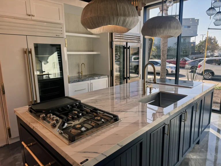 Modern kitchen with a marble island, built-in gas cooktop, sink, gold fixtures, pendant lights, and large windows showing parked cars and buildings outside.