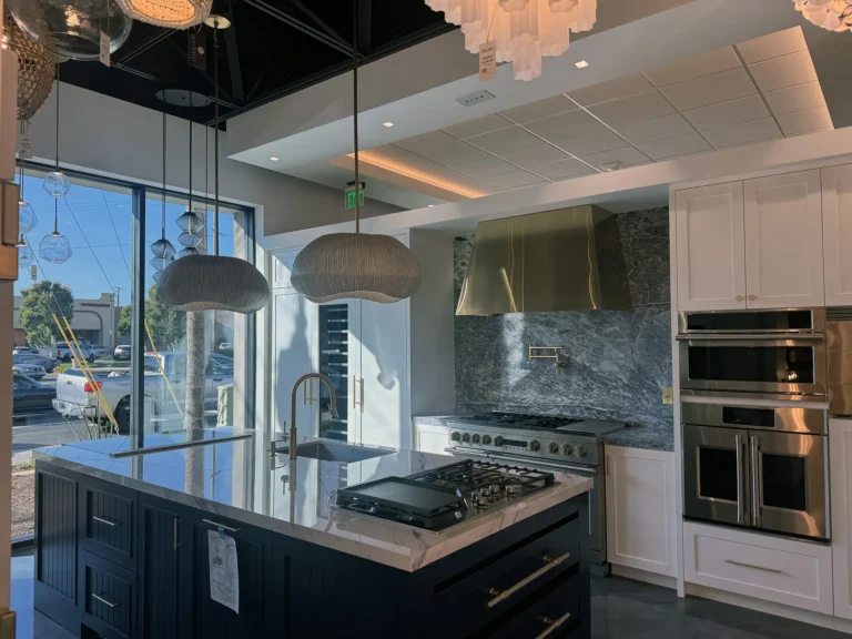 A modern kitchen with a marble island, built-in stove, double oven, gold fixtures, and large windows letting in natural light. Pendant lights hang above the island and a metallic range hood is above the stove.