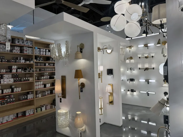 A lighting store interior with white display walls featuring various wall lamps and light fixtures, shelves of light bulbs on the left, and ceiling fans and pendant lights overhead on a glossy floor.