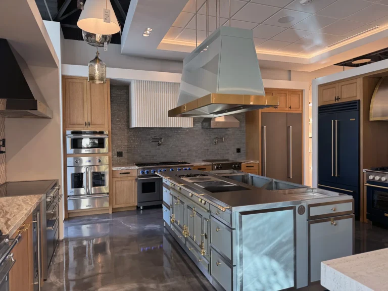 Modern kitchen showroom with stainless steel appliances, light wood cabinets, a large blue and brass island stove, gray backsplash, marble countertops, and overhead range hoods under bright ceiling lights.