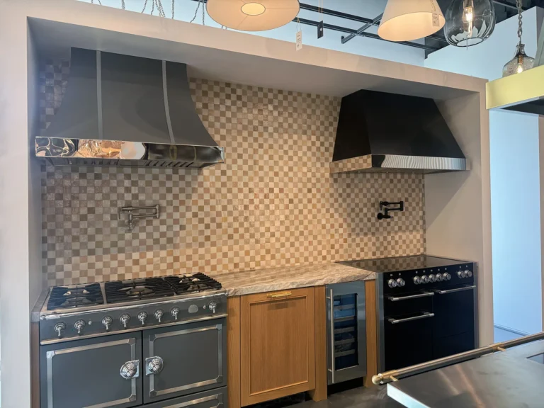 A modern kitchen display features two stovetops with metal range hoods, a marble countertop, a wooden cabinet with a built-in wine cooler, and a beige checkered tile backsplash under pendant lights.