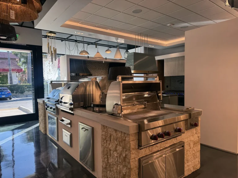 A modern indoor kitchen showroom displays various stainless steel grills and cooking appliances on stone countertops, with pendant lights above and sunlight coming through large windows.