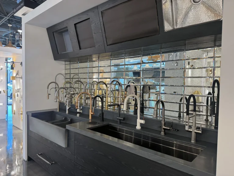 A showroom display of various modern kitchen faucets installed on a long black countertop with three sinks, set against a mirrored tile backsplash.