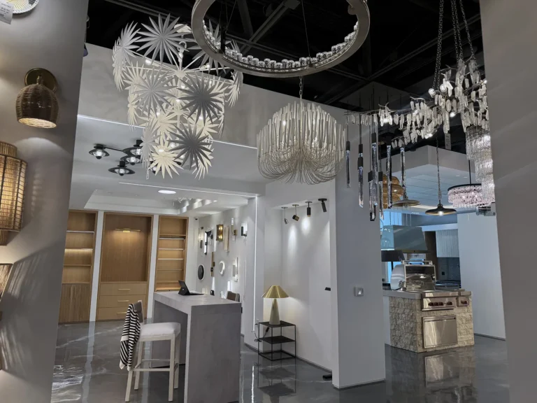 A modern showroom with a glossy floor, displaying various stylish light fixtures including chandeliers and pendant lamps, with a reception desk, stools, wall decor, and wooden shelves in the background.