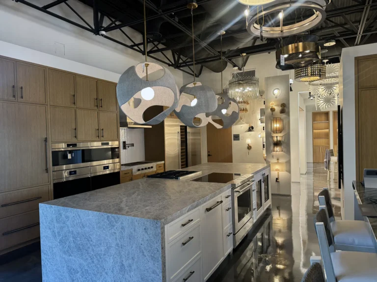 Modern kitchen showroom featuring a marble island with built-in appliances, wooden cabinets, and unique pendant and ceiling lights. The space has a sleek, contemporary design with glossy floors and stylish decor.