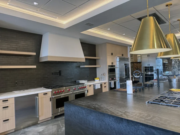 Modern kitchen with light wood cabinets, black countertops, stainless steel appliances, a large stove, built-in oven, and gold pendant lights hanging above a central island with a built-in gas cooktop.