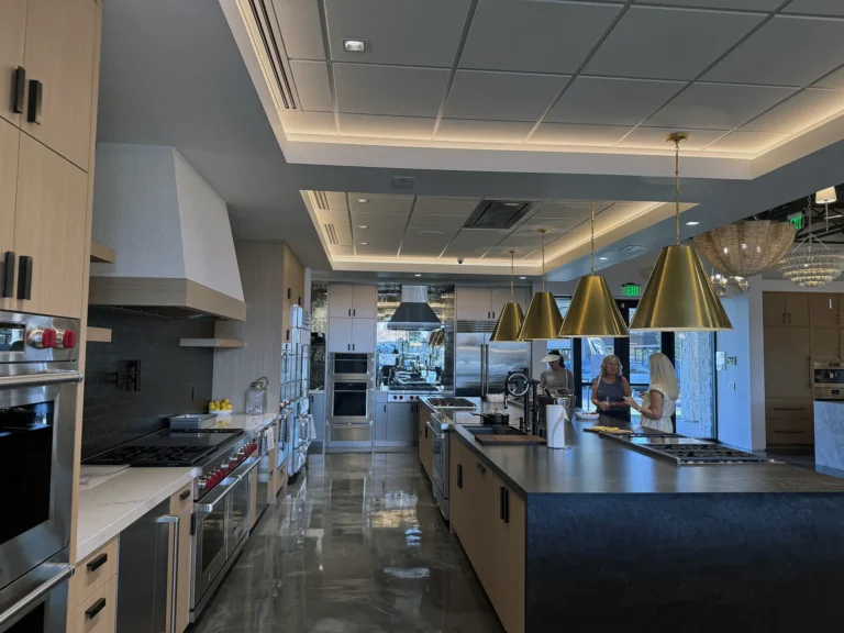 A modern, spacious kitchen showroom with wooden cabinets, stainless steel appliances, large island, gold pendant lights, and two women talking near the windowed end of the room. The floor is shiny and reflective.