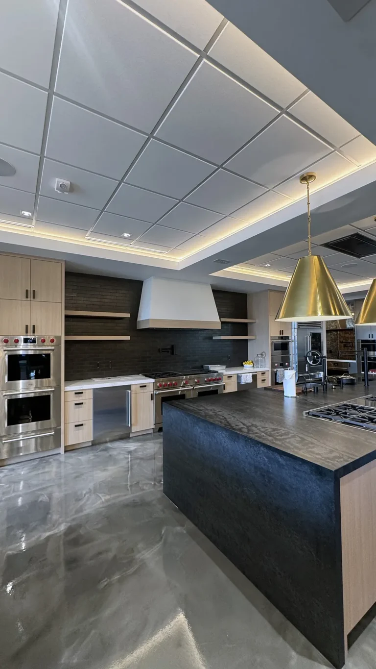 Modern kitchen with light wood cabinets, black countertops, double ovens, a large island with a gas cooktop, and two gold pendant lights. The floor is polished concrete and the ceiling has recessed lighting.
