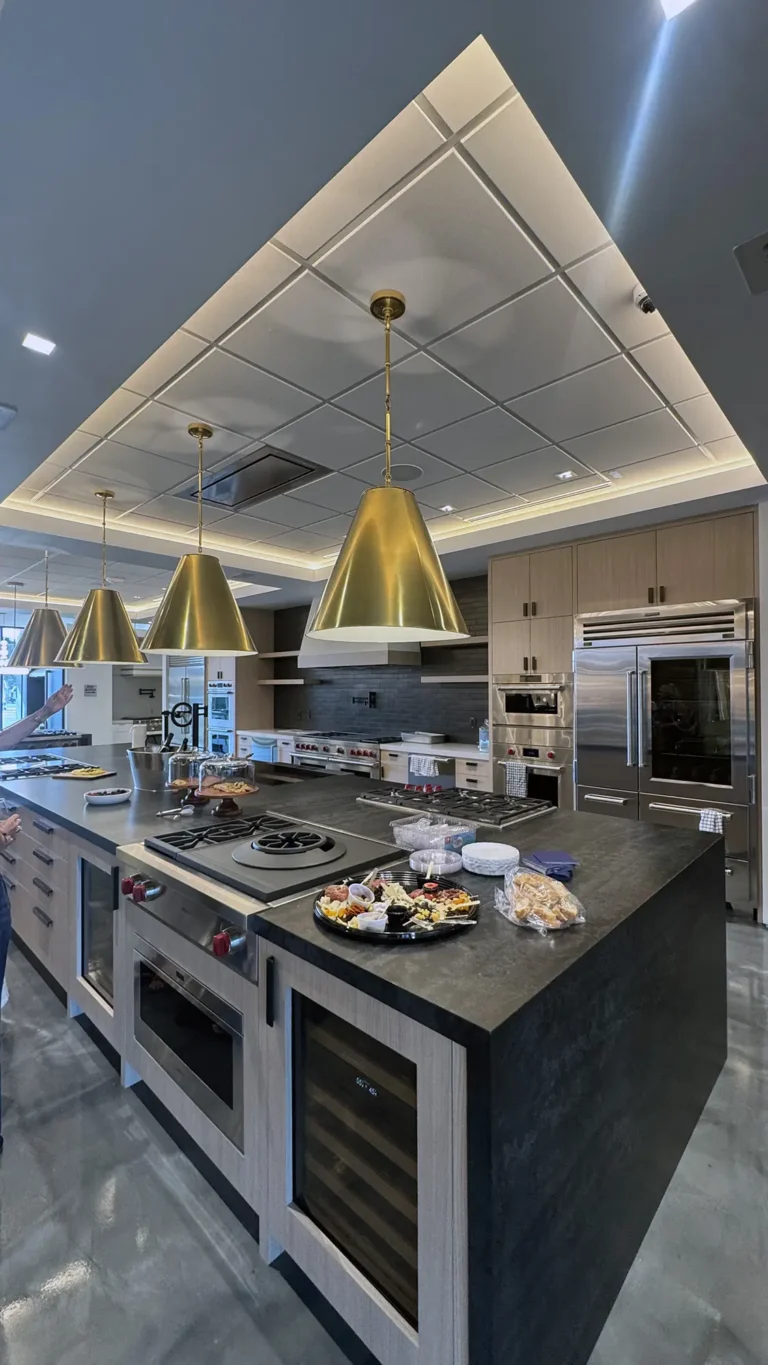 A modern kitchen with wood cabinets, stainless steel appliances, three large gold pendant lights, and a dark island countertop displaying assorted snacks and pastries. The floor is polished gray concrete.