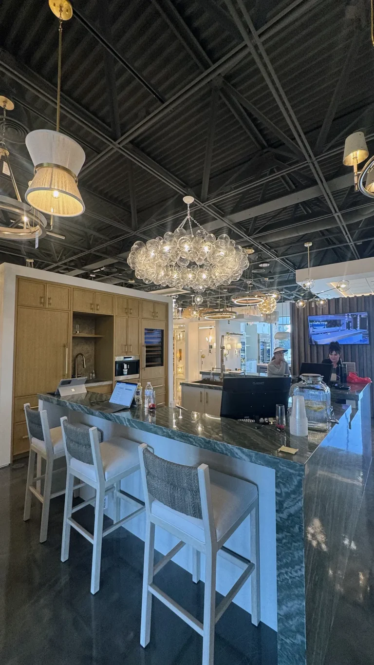A modern kitchen showroom with a marble island, white bar stools, elegant light fixtures, and two people working at a computer near a coffee station under a high, exposed ceiling.