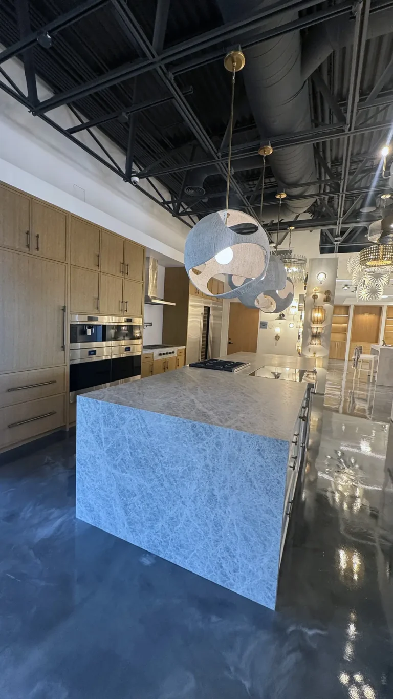Modern kitchen showroom with a large marble island, built-in wooden cabinets, stainless steel appliances, and unique spherical pendant lights hanging from an exposed black ceiling. The floor is shiny and reflective.