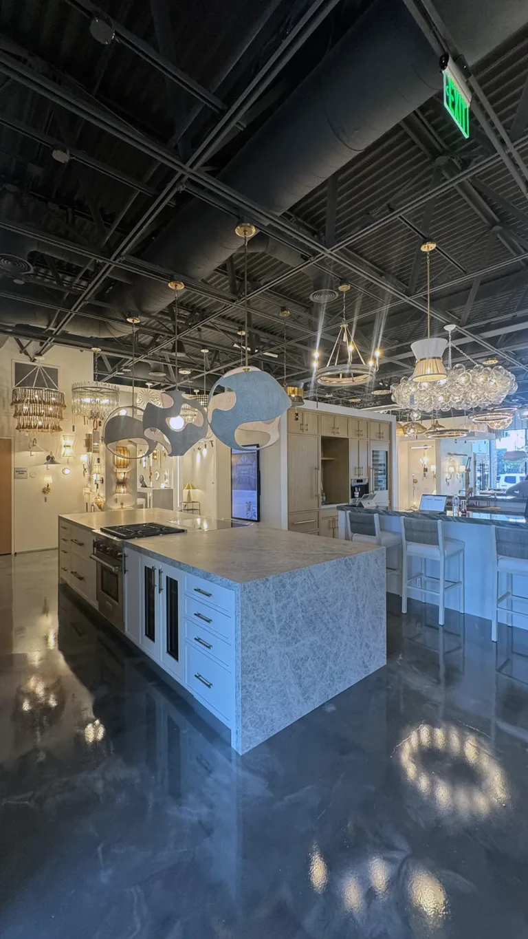 A modern showroom kitchen features a large marble island, white cabinetry, elegant pendant lights, bar stools, and shiny polished floors, with various lighting fixtures displayed in the background.