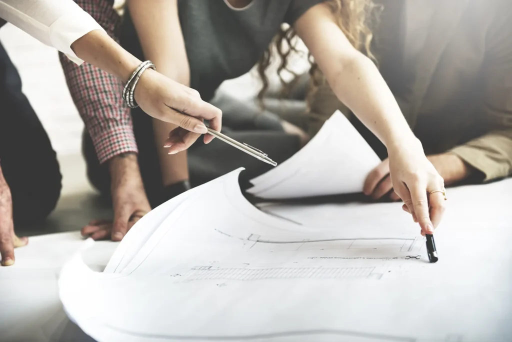Three people review and point to details on large architectural blueprints spread out on a table, using pens and hands to highlight sections of the plans, as they discuss options for architectural lighting design services.