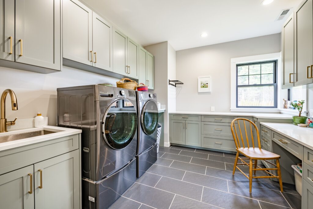 Modern laundry room with gray cabinets, black front-loading washer and dryer, a utility sink, built-in countertops, a wooden chair, and a large window letting in natural light.