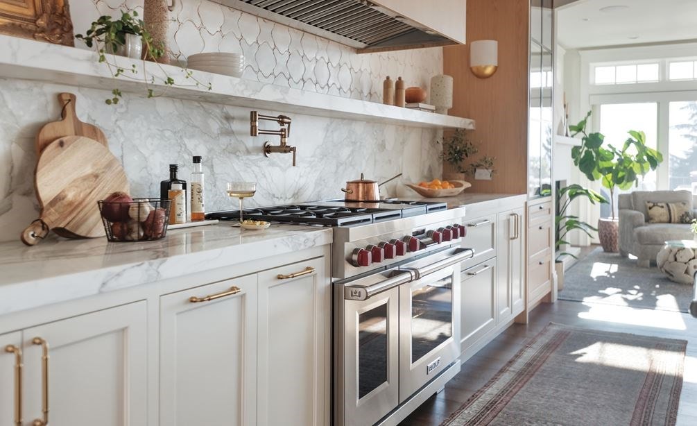 Bright, modern kitchen with white cabinets, gold hardware, marble backsplash and countertops, stainless steel stove with red knobs, open shelving, potted plants, and natural light streaming through large windows.